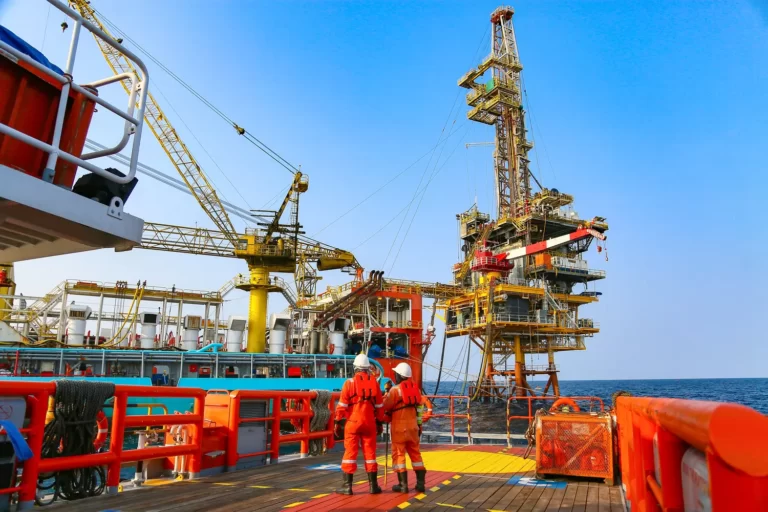Two safety-geared workers on a vessel deck looking toward a large offshore oil or gas production platform against a blue sky.