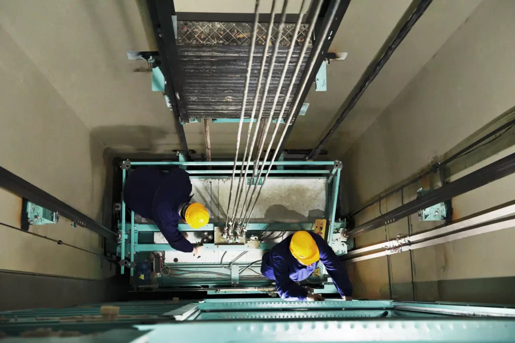 Aerial view looking down an elevator shaft, showing two technicians in safety gear working on the top of the lift car during maintenance.