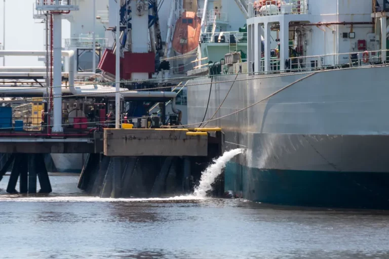 Large commercial vessel moored at a pier discharging a strong stream of water from its hull, representing a ballast water management operation.